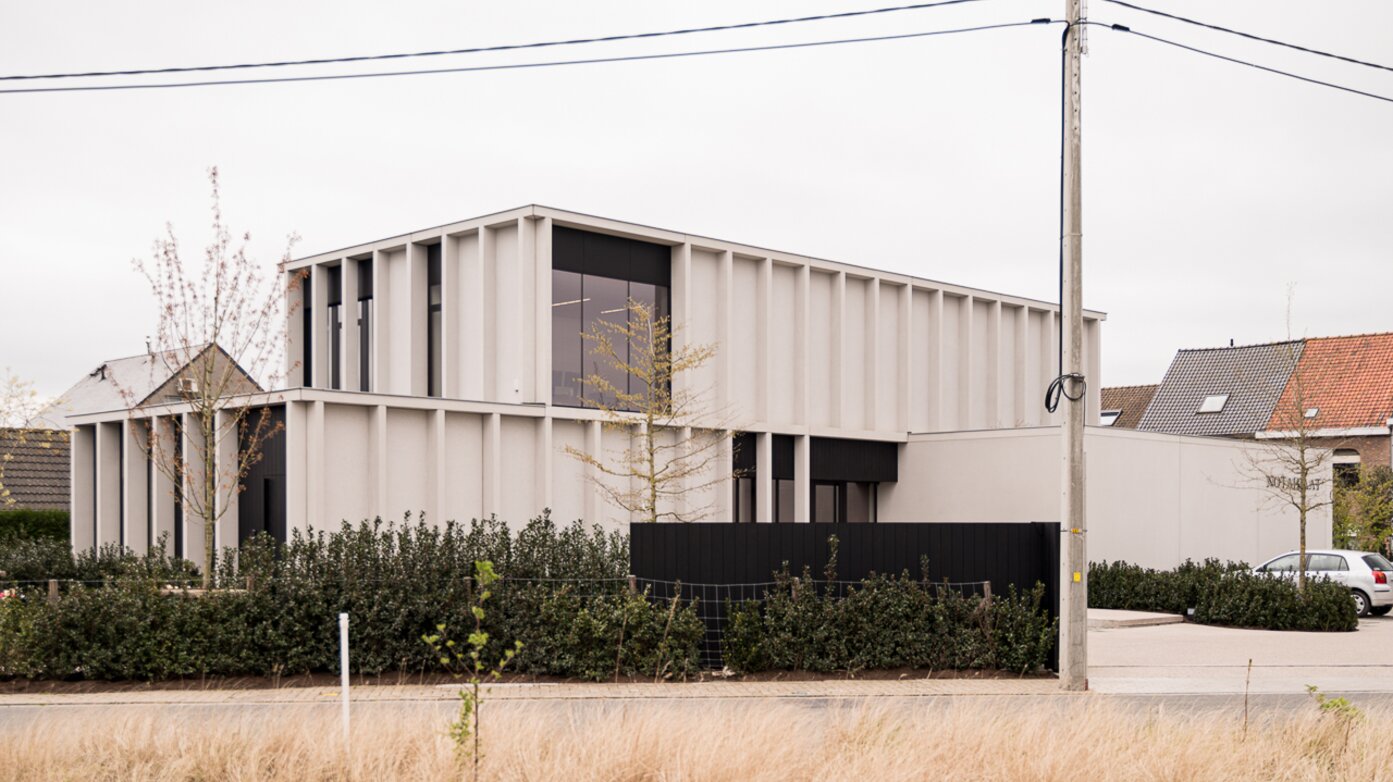 Straatzicht van het notariaat in Meulebeke, ontworpen door GC Architecten: een licht gekleurd volume met verticale gevellamellen, donkere raamkaders en een lagere zijvleugel, omgeven door een strakke haag en siergrassen.