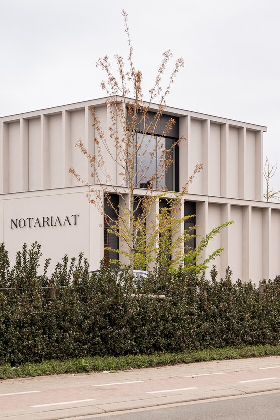 Straatzicht van het notariaat in Meulebeke, ontworpen door GC Architecten: een licht gekleurd volume met verticale gevellamellen, donkere raamkaders en een lagere zijvleugel, omgeven door een strakke haag en siergrassen.
