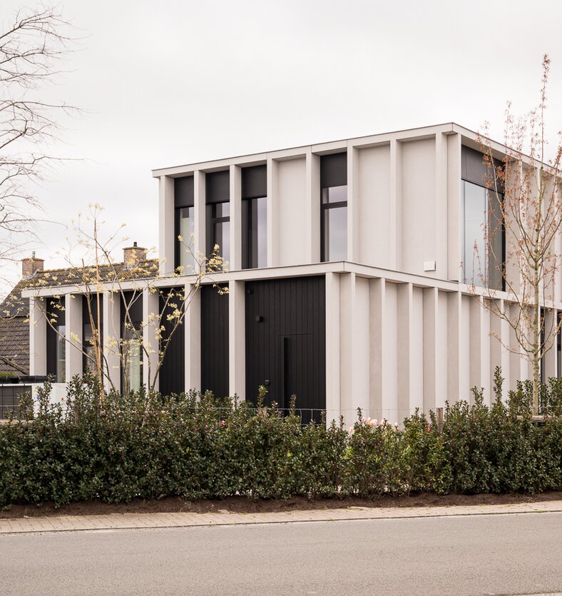 Straatzicht van het notariaat in Meulebeke, ontworpen door GC Architecten: een licht gekleurd volume met verticale gevellamellen, donkere raamkaders en een lagere zijvleugel, omgeven door een strakke haag en siergrassen.