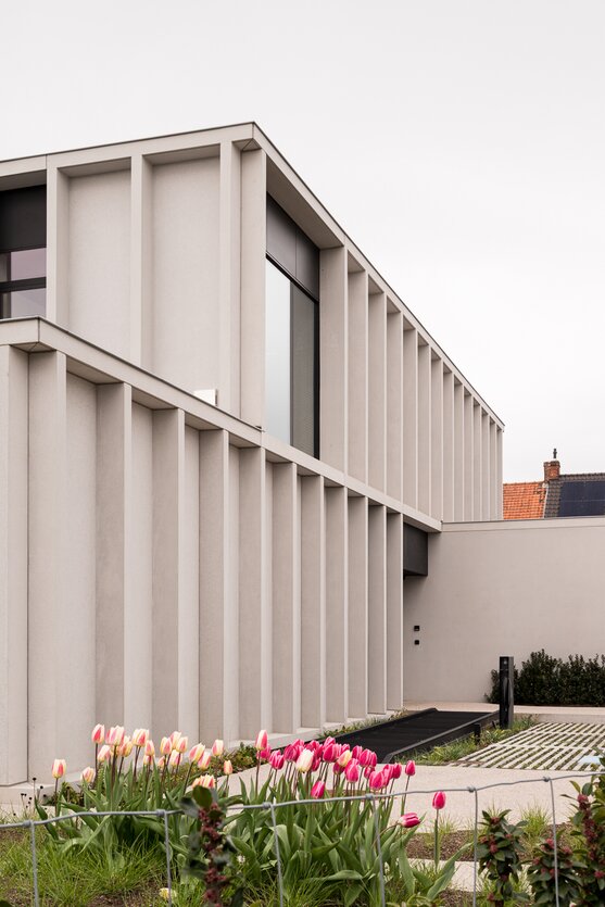 Straatzicht van het notariaat in Meulebeke, ontworpen door GC Architecten: een licht gekleurd volume met verticale gevellamellen, donkere raamkaders en een lagere zijvleugel, omgeven door een strakke haag en siergrassen.