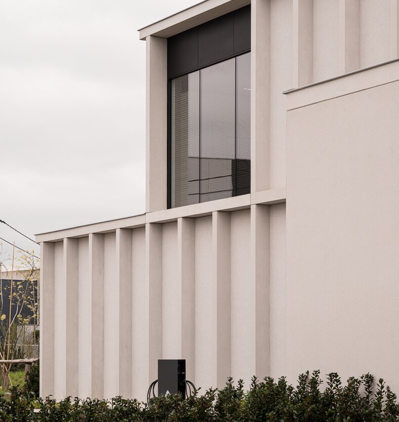 Straatzicht van het notariaat in Meulebeke, ontworpen door GC Architecten: een licht gekleurd volume met verticale gevellamellen, donkere raamkaders en een lagere zijvleugel, omgeven door een strakke haag en siergrassen.