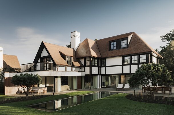 Tuinzicht op een karaktervolle villa in Knokke door GC Architecten: witte gevels met donker vakwerk, rode pannendaken met meerdere dakkapellen, een balkon op de eerste verdieping en grote glaspartijen op het gelijkvloers die uitgeven op een terras met zwemvijver en gesnoeide dennen. | © Bert Demasure