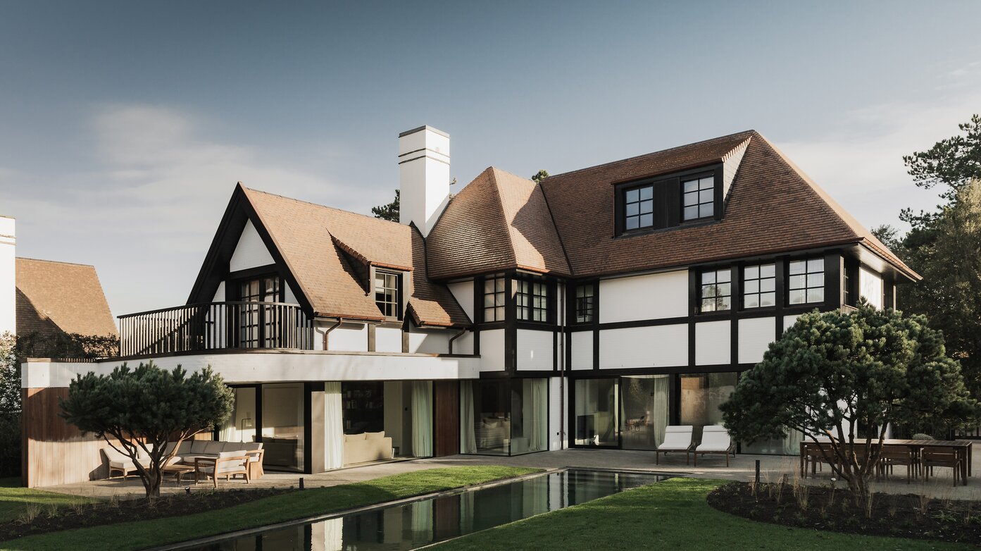 Tuinzicht op een karaktervolle villa in Knokke door GC Architecten: witte gevels met donker vakwerk, rode pannendaken met meerdere dakkapellen, een balkon op de eerste verdieping en grote glaspartijen op het gelijkvloers die uitgeven op een terras met zwemvijver en gesnoeide dennen. | © Bert Demasure