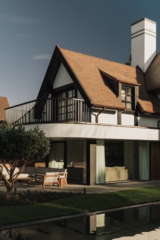 Tuinzicht op een karaktervolle villa in Knokke door GC Architecten: witte gevels met donker vakwerk, rode pannendaken met meerdere dakkapellen, een balkon op de eerste verdieping en grote glaspartijen op het gelijkvloers die uitgeven op een terras met zwemvijver en gesnoeide dennen. | © Bert Demasure