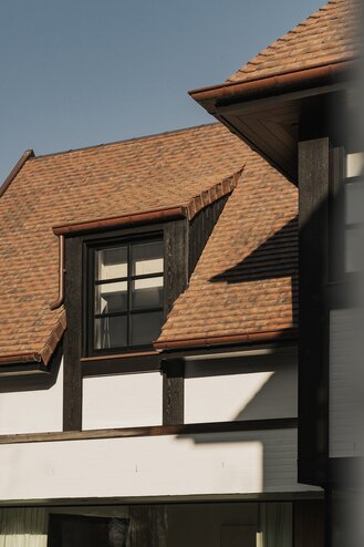 Tuinzicht op een karaktervolle villa in Knokke door GC Architecten: witte gevels met donker vakwerk, rode pannendaken met meerdere dakkapellen, een balkon op de eerste verdieping en grote glaspartijen op het gelijkvloers die uitgeven op een terras met zwemvijver en gesnoeide dennen. | © Bert Demasure