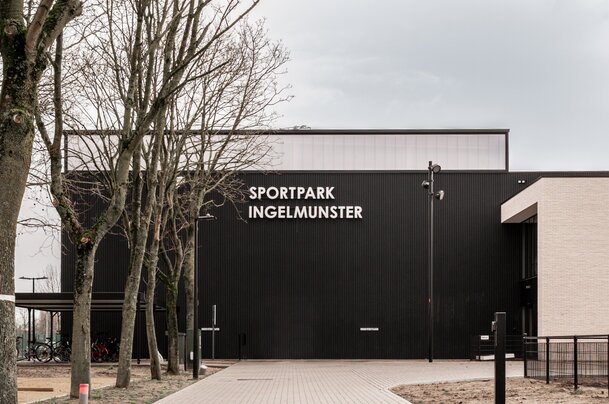 Vooraanzicht van Sportpark Ingelmunster door GC Architecten: een hoog volume met zwarte verticaal geribde gevelbekleding en witte letters, bereikbaar via een bestrate toegangsweg in halfsteens verband, geflankeerd door bestaande bomen en een overdekte fietsenstalling.
