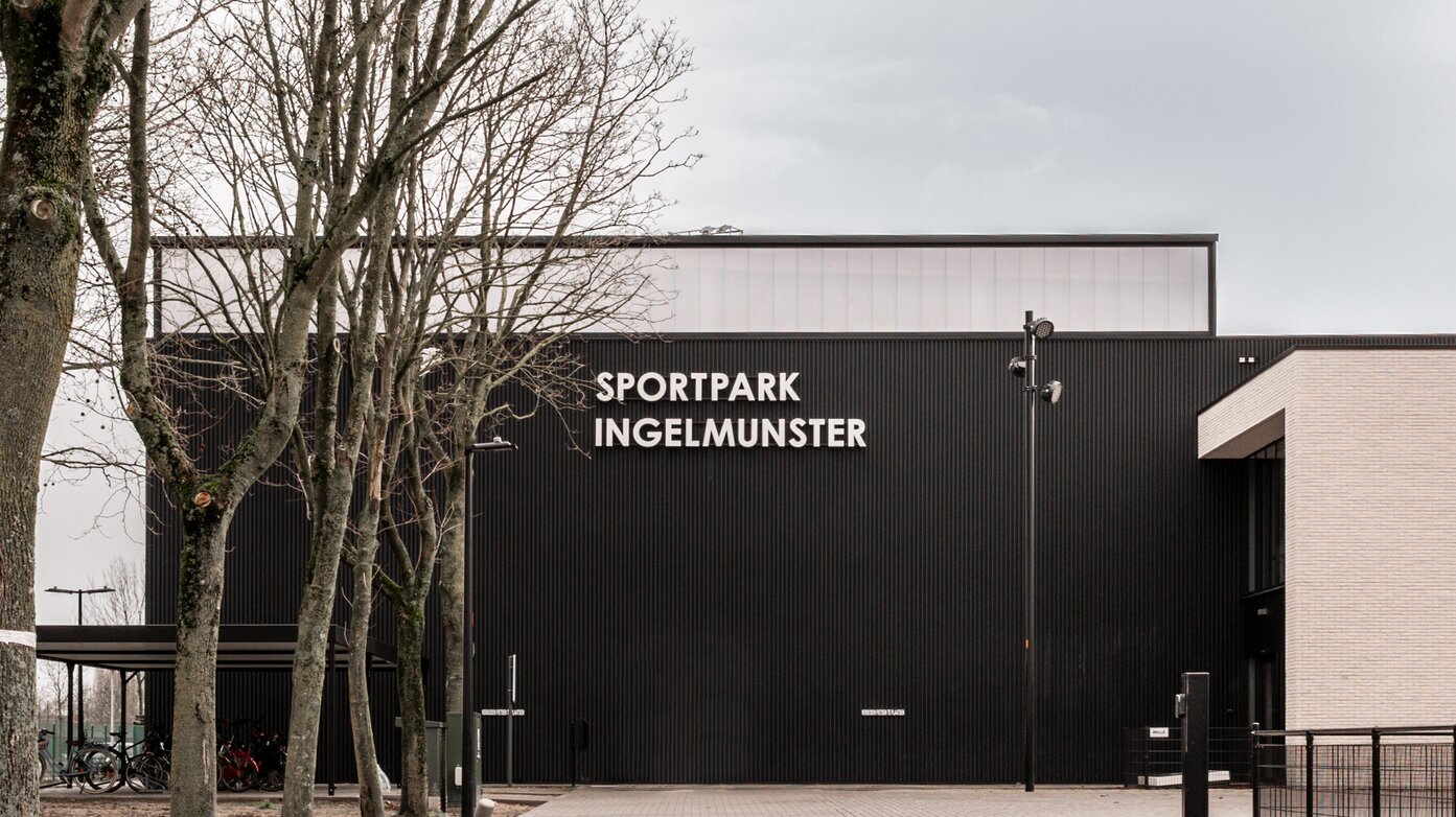 Vooraanzicht van Sportpark Ingelmunster door GC Architecten: een hoog volume met zwarte verticaal geribde gevelbekleding en witte letters, bereikbaar via een bestrate toegangsweg in halfsteens verband, geflankeerd door bestaande bomen en een overdekte fietsenstalling.