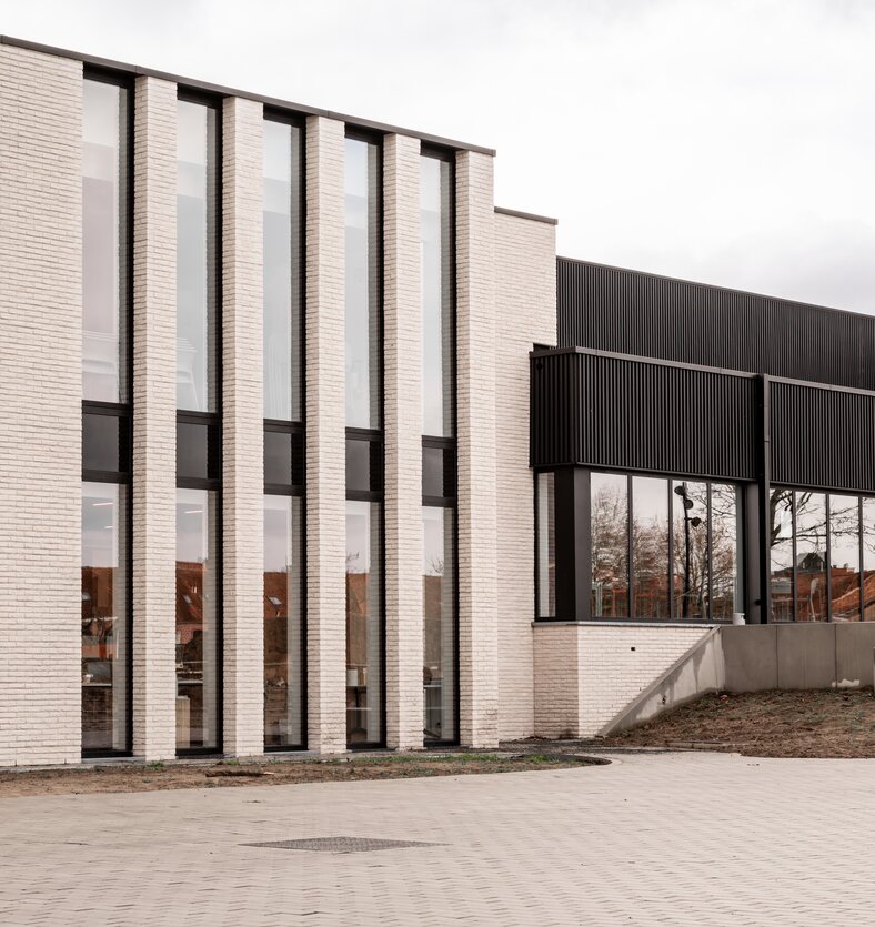 Zijaanzicht van Sportpark Ingelmunster door GC Architecten: een compact volume in lichte gevelsteen met slanke, hoge raampartijen, aansluitend op de zwarte sporthal, gezien vanaf het ruime bestrate voorplein.
