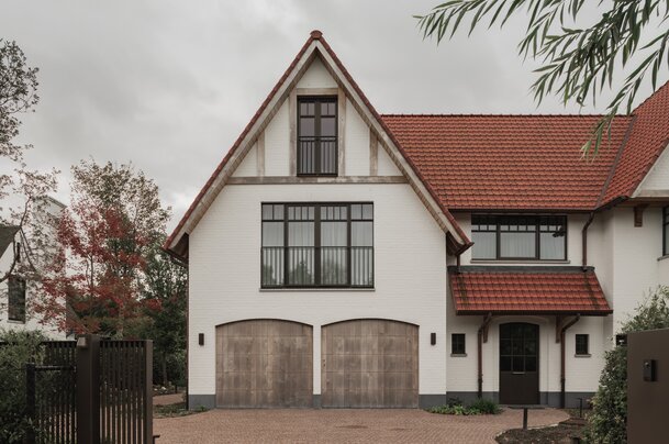 Voorgevel van een cottagevilla in Knokke door GC Architecten: een witgeschilderde bakstenen gevel met puntgevel, houten balken in de topgevel, twee rondbogige houten garagepoorten en donkere raampartijen met fijne roedeverdeling. | © Bert Demasure