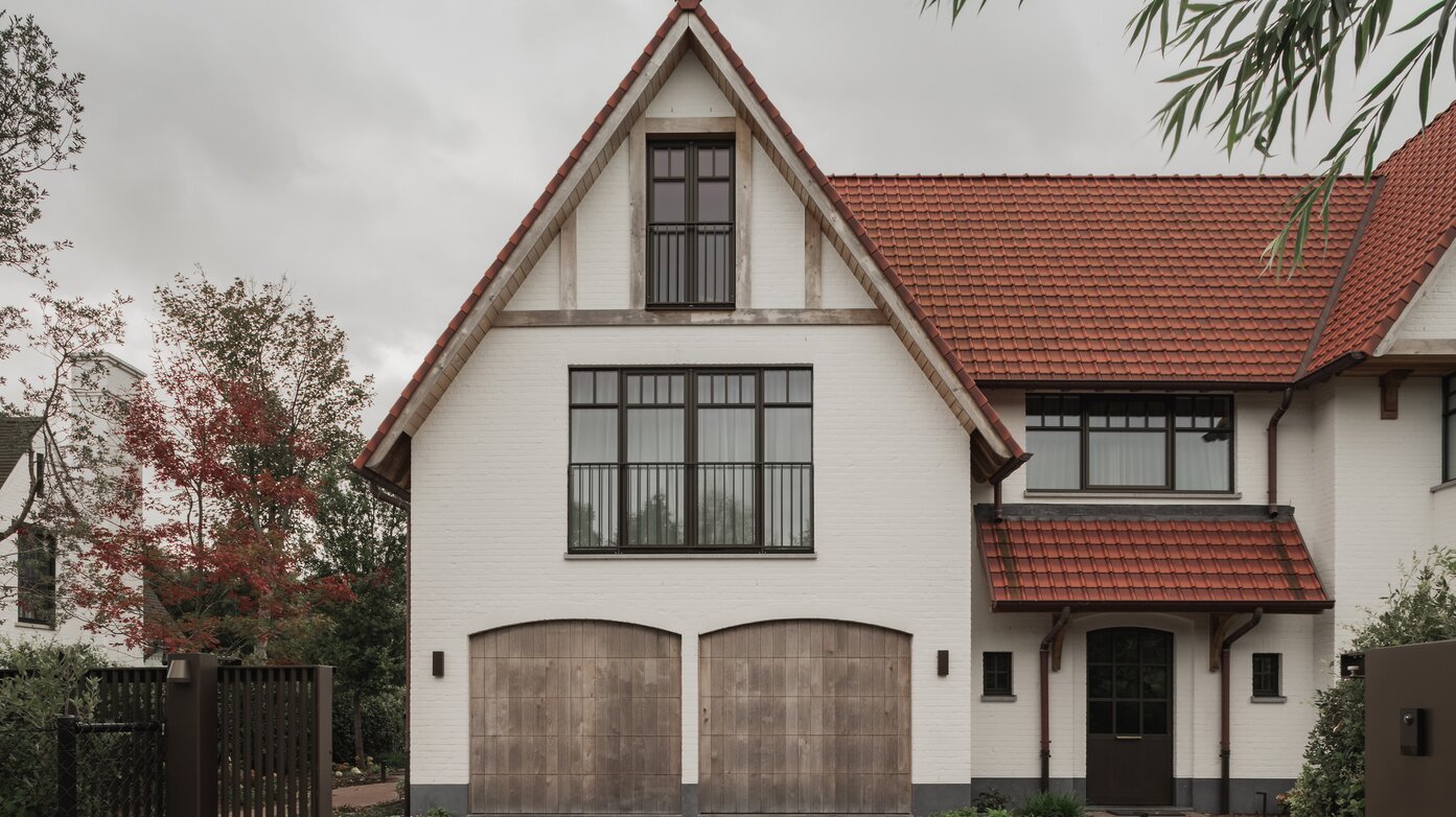 Voorgevel van een cottagevilla in Knokke door GC Architecten: een witgeschilderde bakstenen gevel met puntgevel, houten balken in de topgevel, twee rondbogige houten garagepoorten en donkere raampartijen met fijne roedeverdeling. | © Bert Demasure