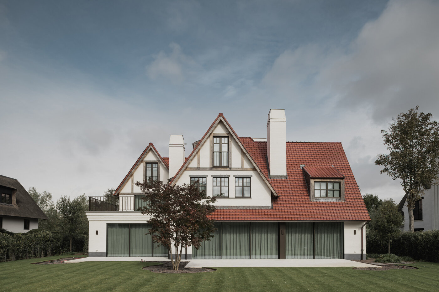 Tuinzicht op een klassieke cottagevilla in Knokke door GC Architecten: witgeschilderde bakstenen gevels met puntgevels, rode pannendaken, witte gemetselde schoorstenen en grote glaspartijen op het gelijkvloers, uitgevend op een verzorgd gazon. | © Bert Demasure