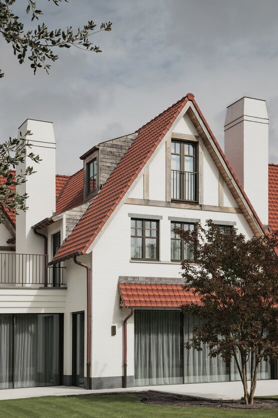 Tuinzicht op een klassieke cottagevilla in Knokke door GC Architecten: witgeschilderde bakstenen gevels met puntgevels, rode pannendaken, witte gemetselde schoorstenen en grote glaspartijen op het gelijkvloers, uitgevend op een verzorgd gazon. | © Bert Demasure