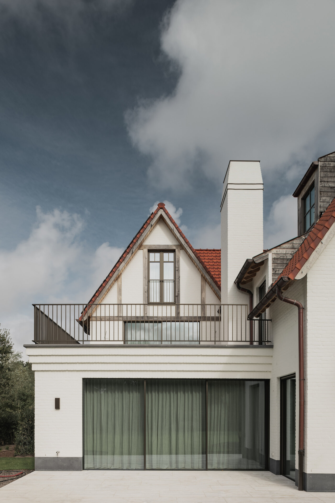 Tuinzicht op een klassieke cottagevilla in Knokke door GC Architecten: witgeschilderde bakstenen gevels met puntgevels, rode pannendaken, witte gemetselde schoorstenen en grote glaspartijen op het gelijkvloers, uitgevend op een verzorgd gazon. | © Bert Demasure