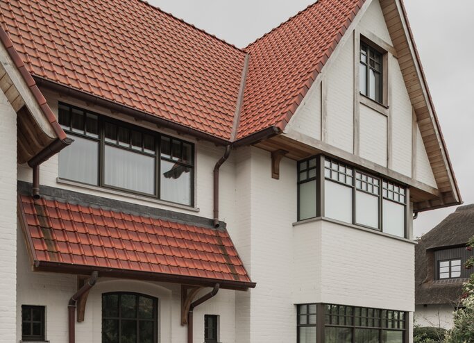 Inkompartij van een cottagevilla in Knokke door GC Architecten: witgeschilderde bakstenen gevel met houten voordeur onder een pannen luifel, geprofileerde houten consoles en een oprit in rode klinkers, omzoomd door een aangelegde voortuin. | © Bert Demasure