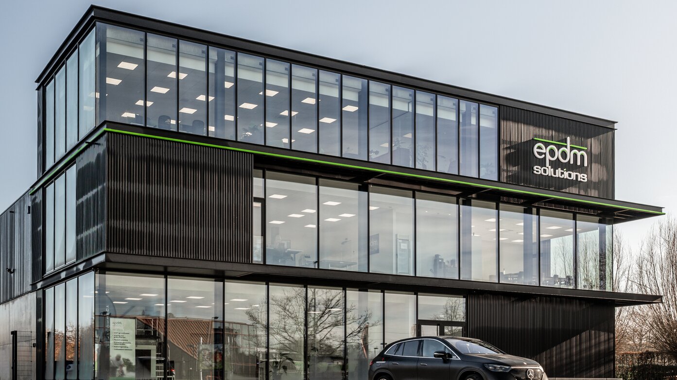 Straatzicht van het kantoor van EPDM Solutions in Roeselare door GC Architecten: een driedelig bedrijfsgebouw met gestapelde volumes in zwarte verticaal geribde gevelbekleding en grote glaspartijen, geaccentueerd door een groene lijn en het bedrijfslogo op de bovenste verdieping.