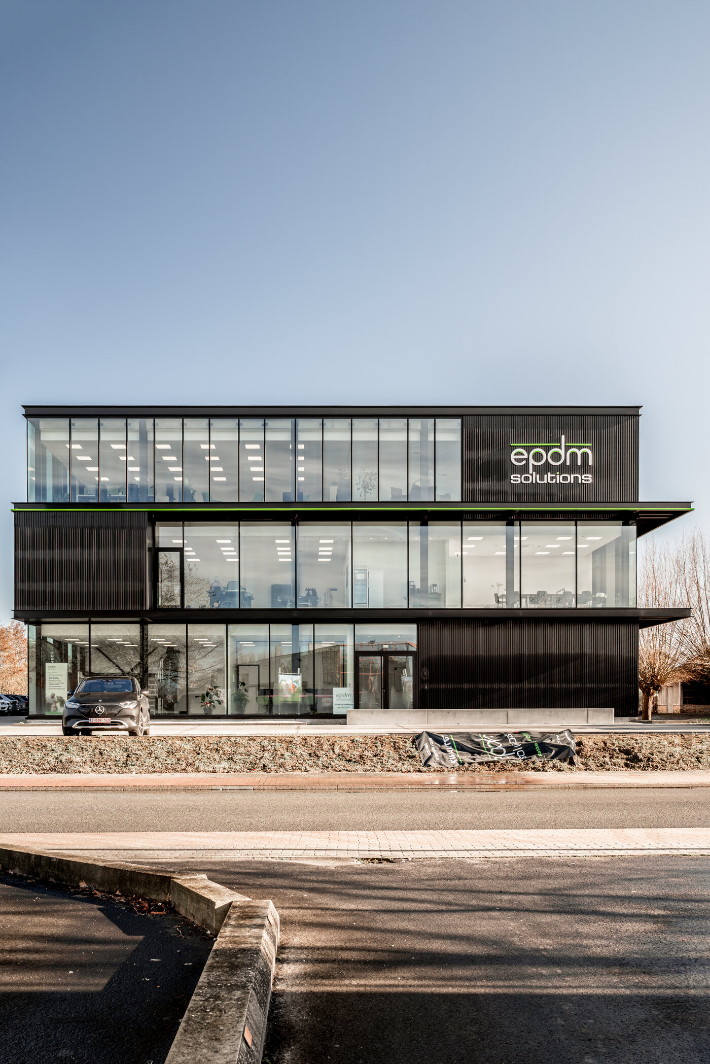 Straatzicht van het kantoor van EPDM Solutions in Roeselare door GC Architecten: een driedelig bedrijfsgebouw met gestapelde volumes in zwarte verticaal geribde gevelbekleding en grote glaspartijen, geaccentueerd door een groene lijn en het bedrijfslogo op de bovenste verdieping.