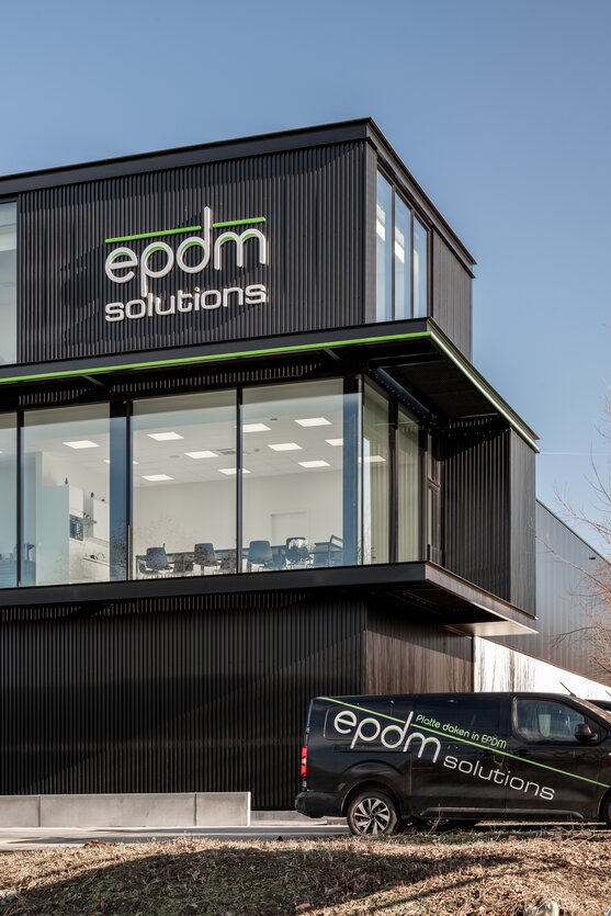 Straatzicht van het kantoor van EPDM Solutions in Roeselare door GC Architecten: een driedelig bedrijfsgebouw met gestapelde volumes in zwarte verticaal geribde gevelbekleding en grote glaspartijen, geaccentueerd door een groene lijn en het bedrijfslogo op de bovenste verdieping.