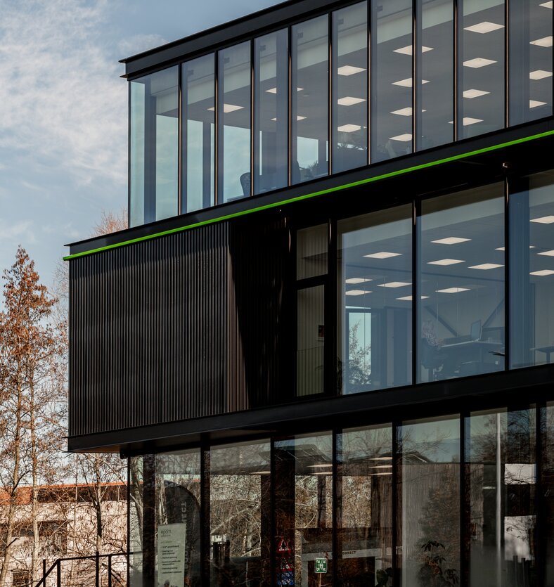 Straatzicht van het kantoor van EPDM Solutions in Roeselare door GC Architecten: een driedelig bedrijfsgebouw met gestapelde volumes in zwarte verticaal geribde gevelbekleding en grote glaspartijen, geaccentueerd door een groene lijn en het bedrijfslogo op de bovenste verdieping.