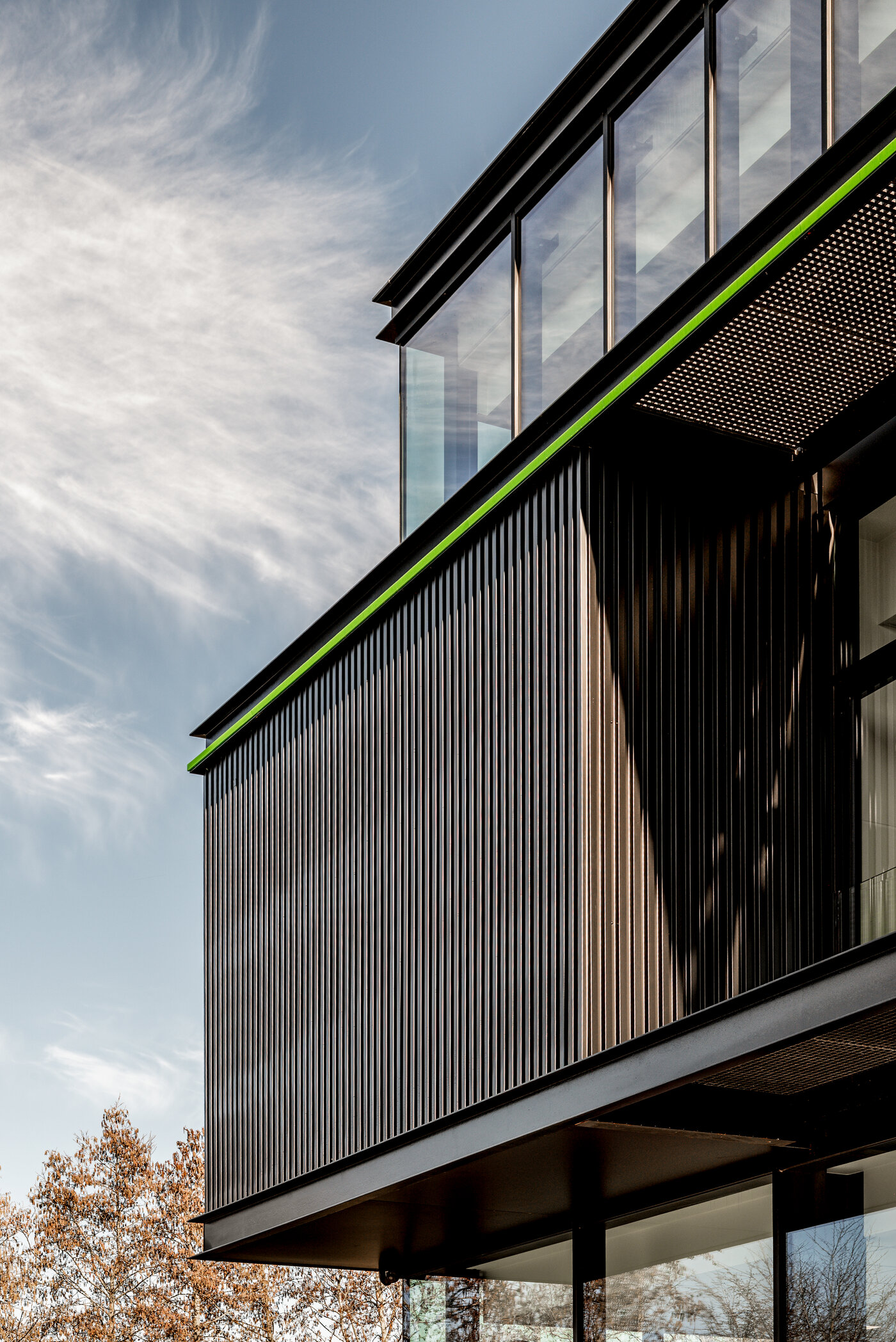 Straatzicht van het kantoor van EPDM Solutions in Roeselare door GC Architecten: een driedelig bedrijfsgebouw met gestapelde volumes in zwarte verticaal geribde gevelbekleding en grote glaspartijen, geaccentueerd door een groene lijn en het bedrijfslogo op de bovenste verdieping.