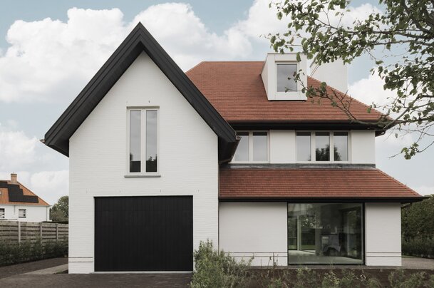 Voorgevel van een hedendaagse cottagevilla in Knokke door GC Architecten: een witgeschilderde bakstenen gevel met steile puntgevel, rode pannendaken, een zwarte garagepoort en een groot glazen venster dat contrasteert met de strakke, witte muurvlakken. | © Bert Demasure