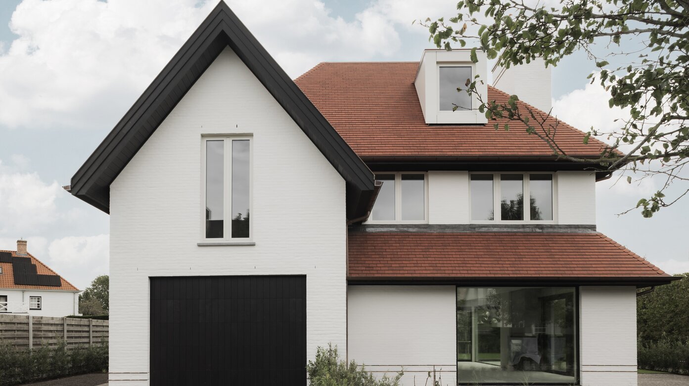 Voorgevel van een hedendaagse cottagevilla in Knokke door GC Architecten: een witgeschilderde bakstenen gevel met steile puntgevel, rode pannendaken, een zwarte garagepoort en een groot glazen venster dat contrasteert met de strakke, witte muurvlakken. | © Bert Demasure
