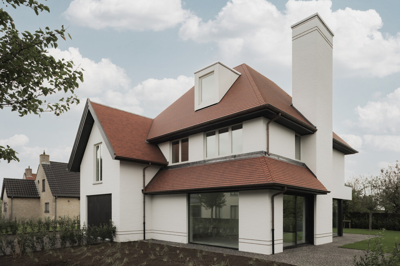 Hoekzicht op een hedendaagse cottagevilla in Knokke door GC Architecten: witgeschilderde bakstenen volumes onder rode pannendaken, een slanke witte schoorsteen en grote frameloze hoekramen op het gelijkvloers die openen naar de tuin. | © Bert Demasure