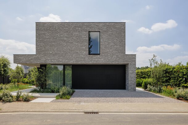 Straatzicht op een kubistische woning met grijze steenstrips, overkragend bovenvolume boven een zwarte garagepoort en een oprit met grind.