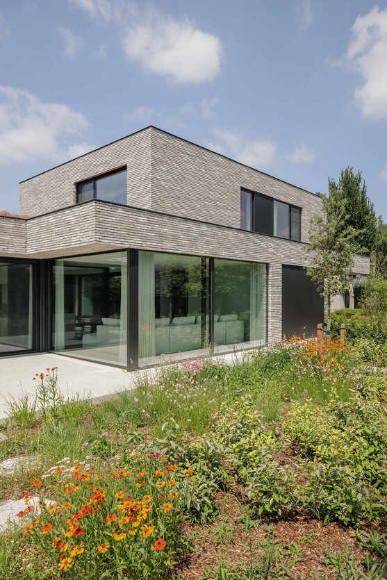 Overstekend bovenvolume in grijze steenstrips boven een glazen hoek met zicht op de woonkamer, omringd door een bloemrijke vasteplantenborder.