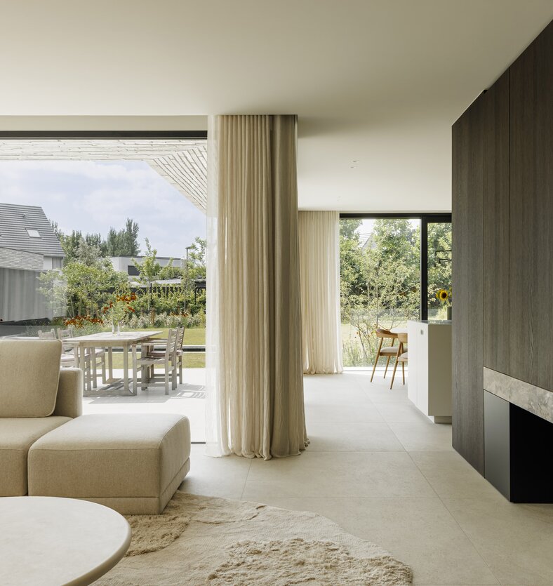 Lichte woonkamer met beige zitbank, gordijnen en een houten haardwand, met doorzicht naar de eethoek, keuken en het terras.