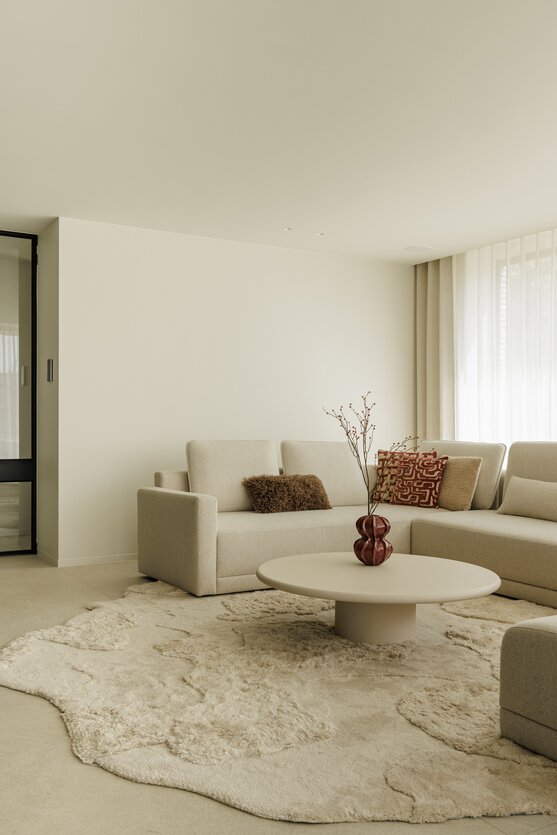 Hoekzetel in beige stof met sierkussens, ronde salontafel met decoratieve vaas en zacht vloerkleed in een lichte woonkamer.