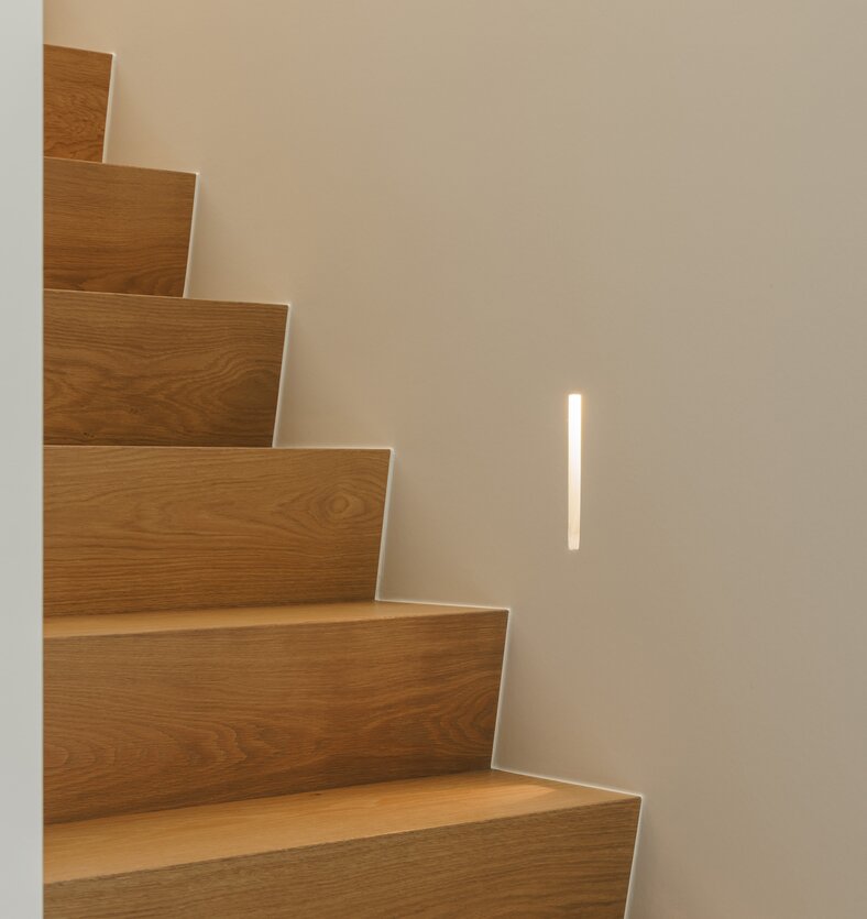 Detail van een houten trap met eiken treden en een verticale inbouwspot in de witte wand.