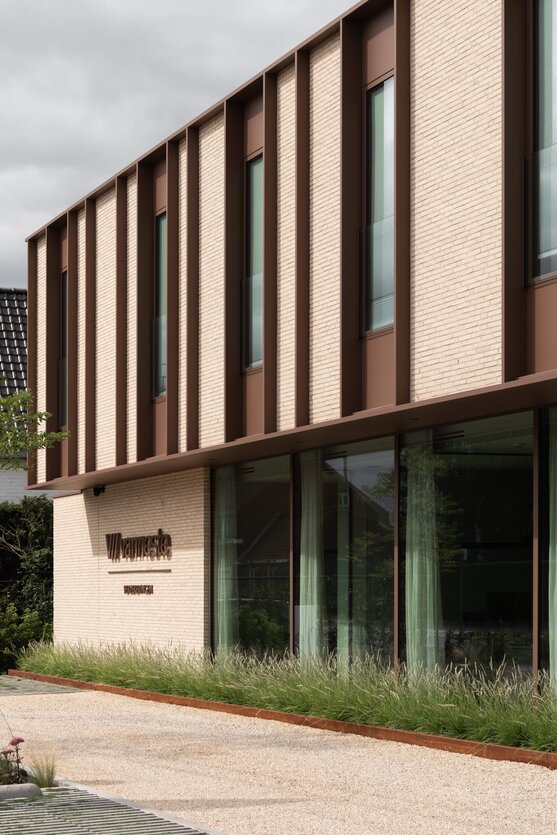 Gevel van een verzekeringskantoor met de bedrijfsnaam in het baksteenmetselwerk verwerkt, verticale gevellamellen en een glazen pui die de omgeving weerspiegelt.
