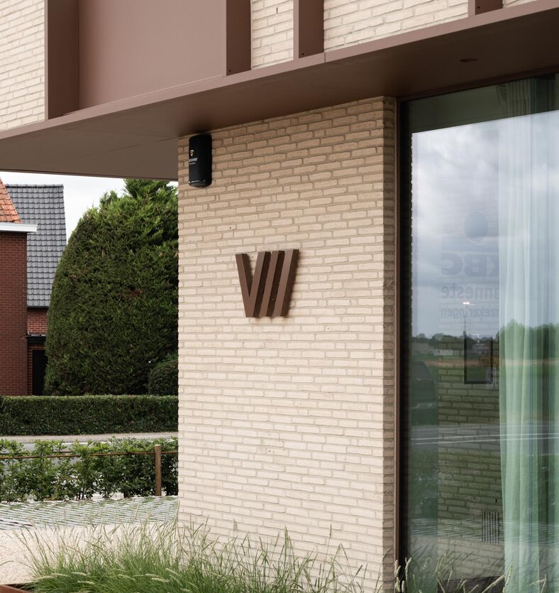 Detailopname van een crèmekleurige baksteenpijler met een bronskleurig wandlogo en glaspartij, ontworpen door GC architecten.