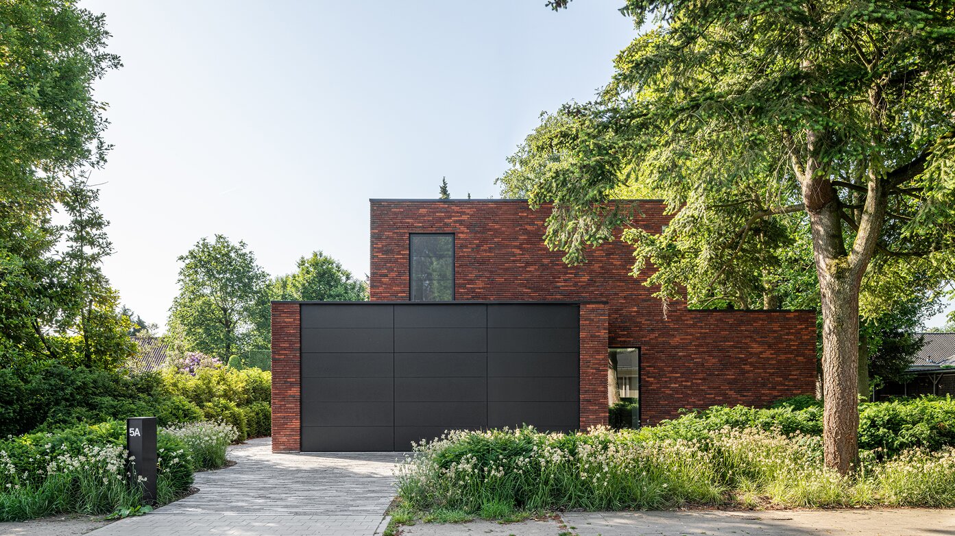 Voorgevel van een hedendaagse villa in donkerrode baksteen met een grote zwarte garagepoort, omgeven door groen en siergrassen.