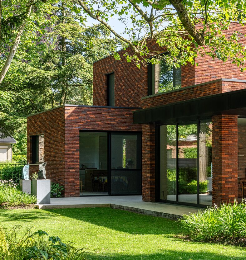 Achtergevel van een bakstenen woning met een glazen tuinkamer die uitgeeft op een terras met sculpturen, ontworpen door GC architecten.